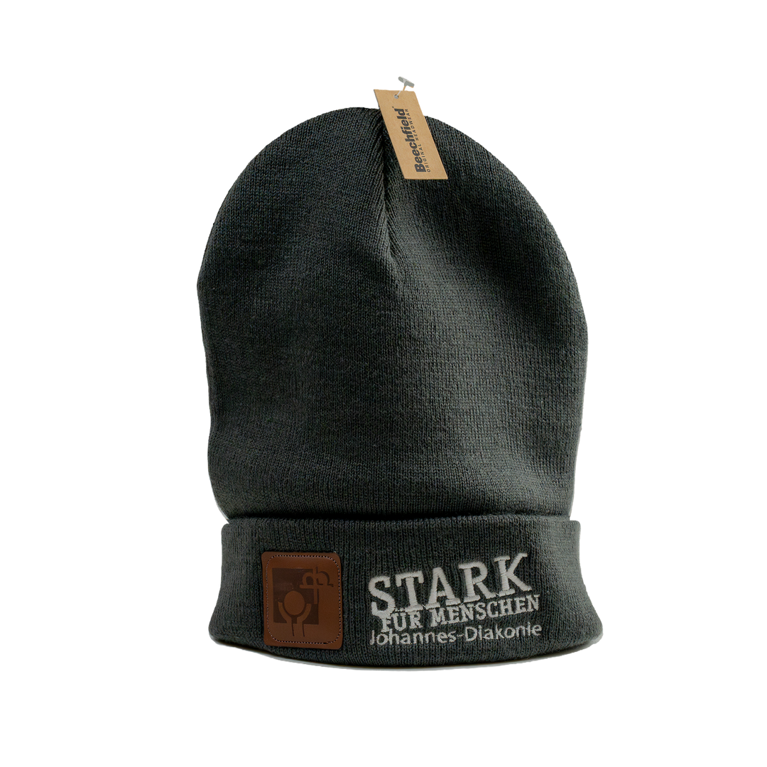 Dunkelgraue gestrickte Beanie-Mütze mit umgeschlagenem Rand und angenähtem Etikett oben. Auf der Vorderseite sind ein braunes Kunstleder-Logo-Patch sowie der weiß gestickte Schriftzug „STARK FÜR MENSCHEN – Johannes-Diakonie“