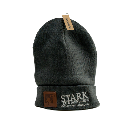 Dunkelgraue gestrickte Beanie-Mütze mit umgeschlagenem Rand und angenähtem Etikett oben. Auf der Vorderseite sind ein braunes Kunstleder-Logo-Patch sowie der weiß gestickte Schriftzug „STARK FÜR MENSCHEN – Johannes-Diakonie“