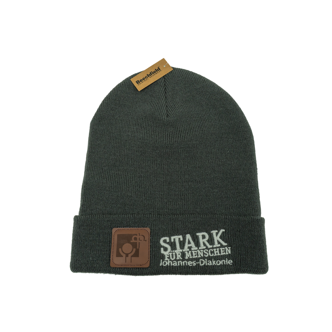 Dunkelgraue gestrickte Beanie-Mütze mit umgeschlagenem Rand. Auf der Vorderseite ist der Schriftzug „STARK FÜR MENSCHEN – Johannes-Diakonie“ in Weiß gestickt sowie ein kleines braunes Logo-Patch aus Kunstleder.