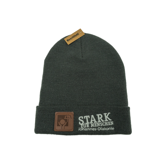 Dunkelgraue gestrickte Beanie-Mütze mit umgeschlagenem Rand. Auf der Vorderseite ist der Schriftzug „STARK FÜR MENSCHEN – Johannes-Diakonie“ in Weiß gestickt sowie ein kleines braunes Logo-Patch aus Kunstleder.
